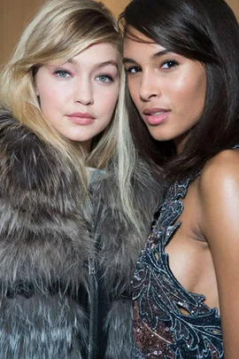 Two women posing closely together, one wearing a fur coat and the other a patterned dress, smiling softly at the camera.