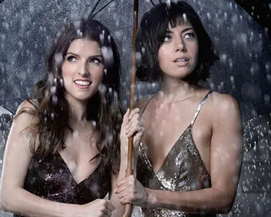 Two women in sparkly dresses stand together under an umbrella in the rain.