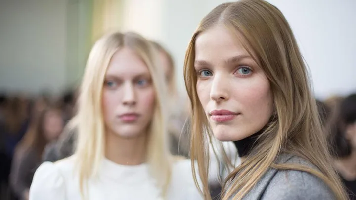 Two women with blonde hair, one in focus wearing a gray top, the other blurred wearing a white top, in a well-lit room.