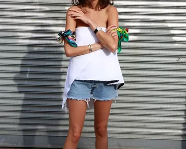 A woman in sunglasses, white top, and denim shorts stands smiling against a metal shutter.