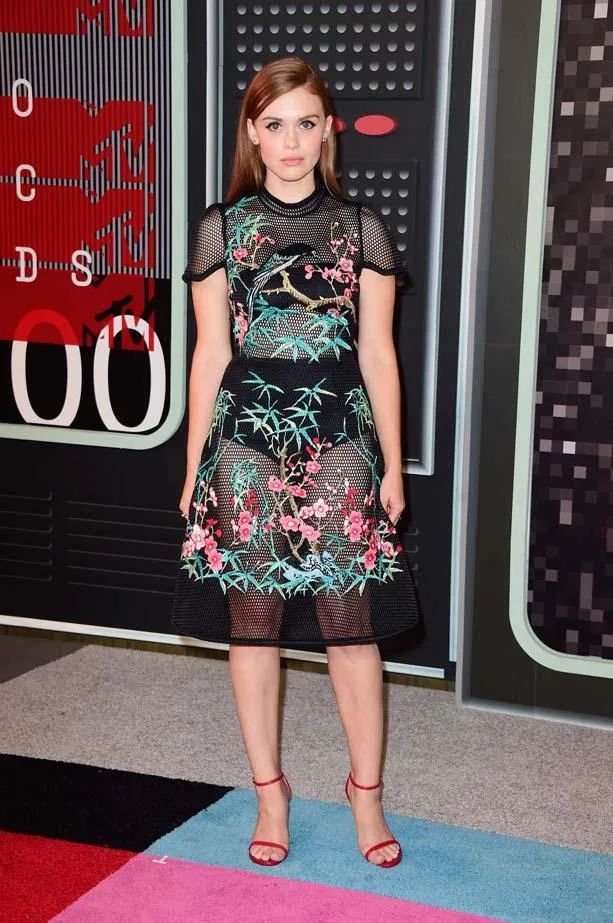 Young woman in a floral dress and red heels on a colorful carpet, standing in front of an "MTV" backdrop.