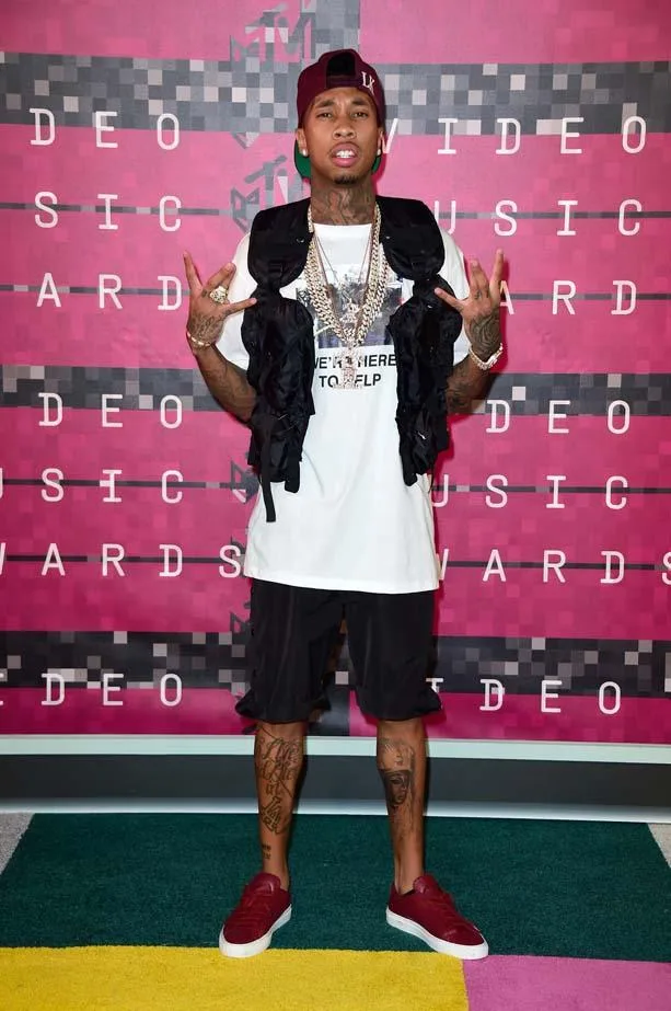 Man posing at MTV Video Music Awards wearing a cap, chains, white shirt, shorts, tattoos, and red sneakers.