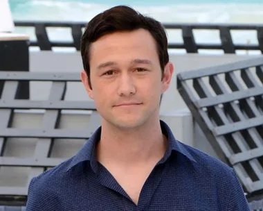 A man in a blue shirt smiling faintly, standing outdoors with a blurred background of metal structures and the sea.