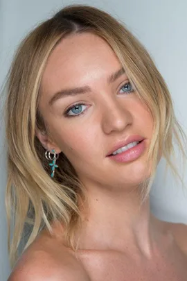 A woman with blonde hair and blue eyes wears a turquoise earring and smiles slightly against a neutral background.