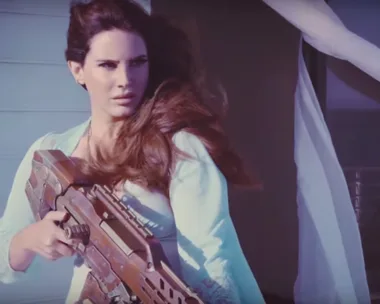 Lana Del Rey holding a large futuristic gun, gazing into the distance, with wind blowing her hair. (From her new music video)