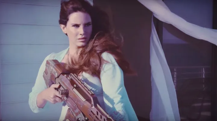 Lana Del Rey holding a large futuristic gun, gazing into the distance, with wind blowing her hair. (From her new music video)