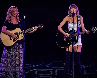 Lisa Kudrow and Taylor Swift play guitars and sing "Smelly Cat" at a concert. Kudrow in patterned skirt, Swift in white dress.