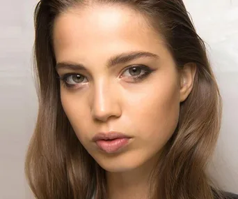 Close-up of a woman's face with natural makeup and long brown hair on a neutral background.