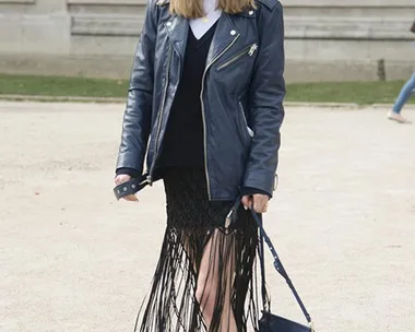 A woman wearing a leather jacket, fringe skirt, and sneakers smiles outdoors, carrying a blue handbag with sunglasses on.