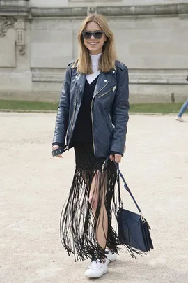 A woman wearing a leather jacket, fringe skirt, and sneakers smiles outdoors, carrying a blue handbag with sunglasses on.