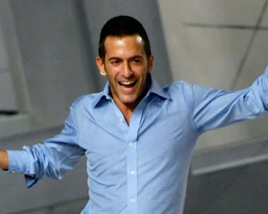 A man with short hair and a light blue shirt is smiling and raising his arms in excitement.