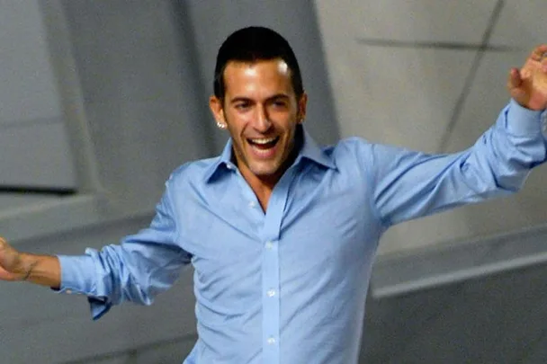 A man with short hair and a light blue shirt is smiling and raising his arms in excitement.