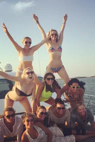 A group of women on a boat, forming a human pyramid, smiling and posing in bikinis, with the sea in the background.