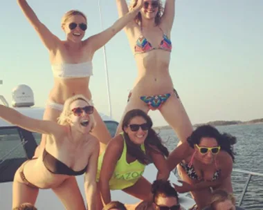 A group of women on a boat, forming a human pyramid, smiling and posing in bikinis, with the sea in the background.