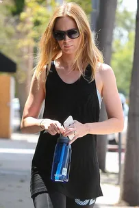 A woman with long hair wearing sunglasses, a black tank top, and holding a water bottle and phone while walking outdoors.