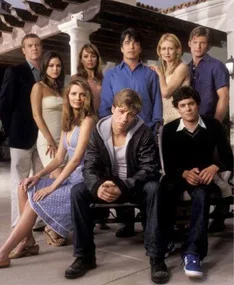 Cast of "The O.C." posing outdoors in front of a stylish house, with nine characters dressed in casual and semi-formal attire.