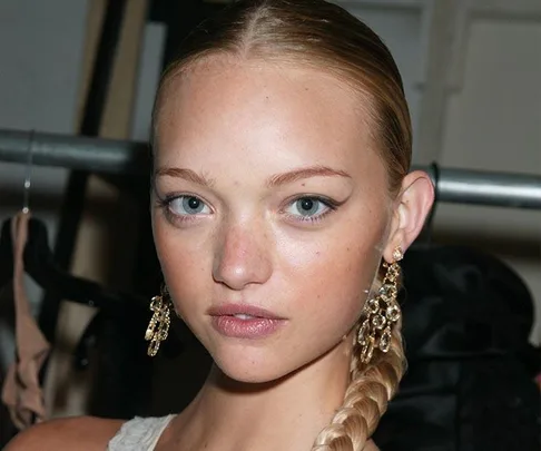 Woman with blonde hair in a braid, wearing large dangling earrings, looking directly at the camera.