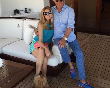 A woman and man sitting on a yacht's deck, both wearing sunglasses, with a small dog nearby.