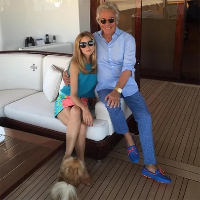 A woman and man sitting on a yacht's deck, both wearing sunglasses, with a small dog nearby.