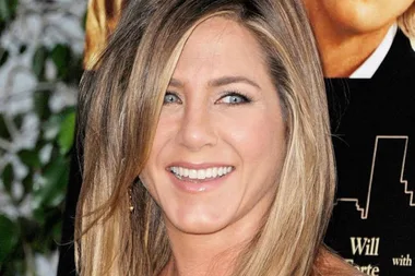 Actress Jennifer Aniston smiling at a public event, with a blurred movie poster in the background.