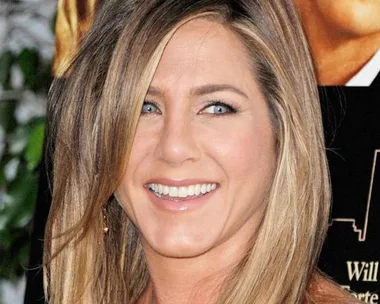 Actress Jennifer Aniston smiling at a public event, with a blurred movie poster in the background.