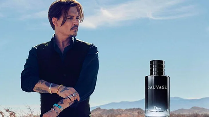 Johnny Depp stands in a desert landscape promoting Dior Sauvage fragrance, with the bottle visible in the foreground.