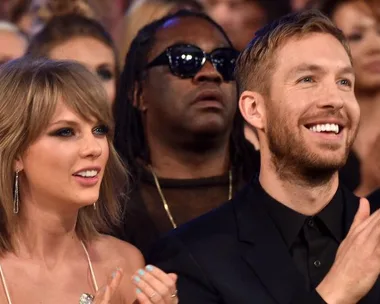 Taylor Swift and Calvin Harris sitting in an audience, smiling and clapping, with other people seated around them.