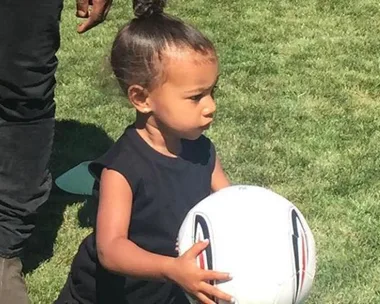 Child in black shirt holding a soccer ball on a grassy field, with part of an adult's leg visible in the background.