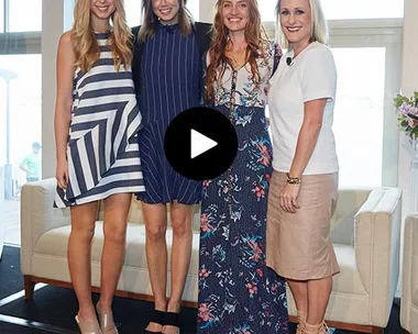 Four women standing side by side in a room with tall windows, dressed in stylish summer dresses and smiling at the camera.