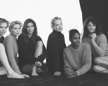 Black and white photo of six women wearing sweaters, seated closely together on the ground, posing for the camera.