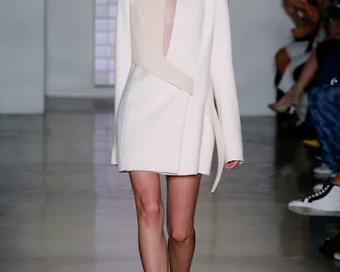 Model in a structured white dress walks the runway at a fashion show, with spectators in the background.