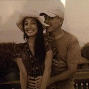 George and Amal Clooney smiling outdoors, Amal wearing a hat, George hugging her from behind.