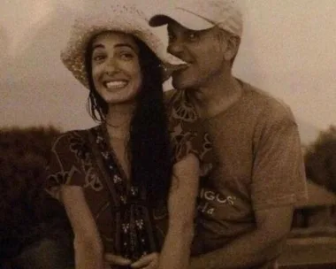 George and Amal Clooney smiling outdoors, Amal wearing a hat, George hugging her from behind.