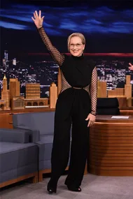 Actress in a black outfit waves on a talk show set with a cityscape backdrop.