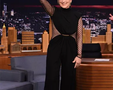 Actress in a black outfit waves on a talk show set with a cityscape backdrop.