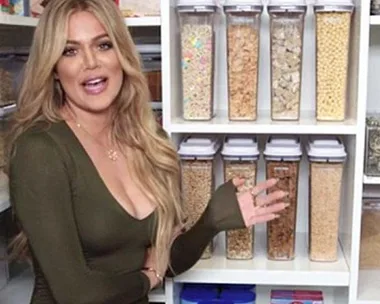 Woman with long blonde hair in olive dress stands in organized pantry with labeled cereal containers.