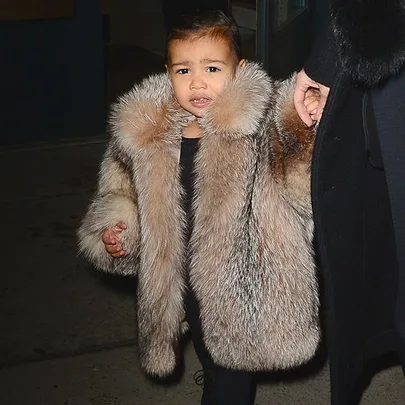 A child wearing a stylish fur coat being escorted by an adult at night.