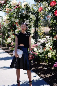 A woman in a black outfit poses in a garden with vibrant roses and a sunny background.