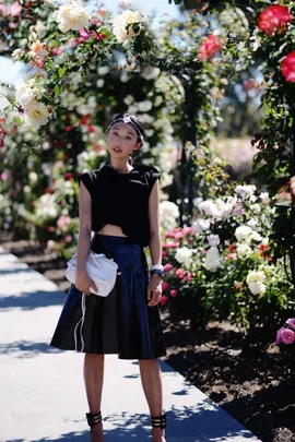 A woman in a black outfit poses in a garden with vibrant roses and a sunny background.