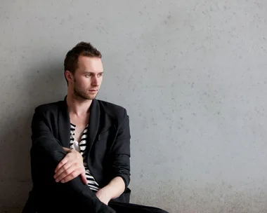 Man wearing black blazer over a striped shirt sits against a plain wall, looking thoughtfully to his left.