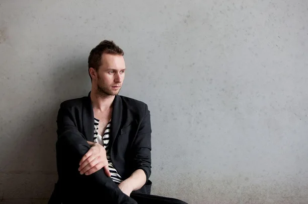 Man wearing black blazer over a striped shirt sits against a plain wall, looking thoughtfully to his left.