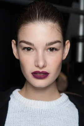 A model with slicked-back hair and bold plum lipstick wearing a white sweater, showcasing a beauty trend.