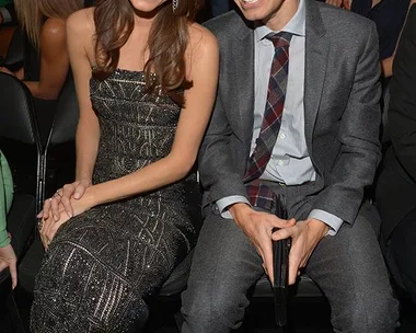 Allison Williams in a glittery dress with a man in a grey suit, sitting together and smiling at an indoor event.