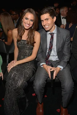 Allison Williams in a glittery dress with a man in a grey suit, sitting together and smiling at an indoor event.