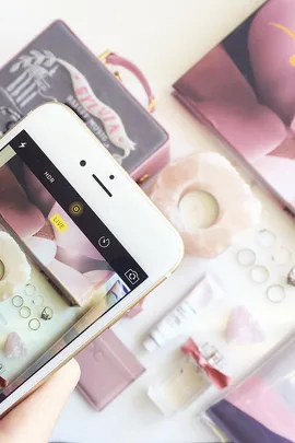 Flatlay of pink-themed accessories including rings, a candle, and perfume, with a smartphone camera capturing the scene.