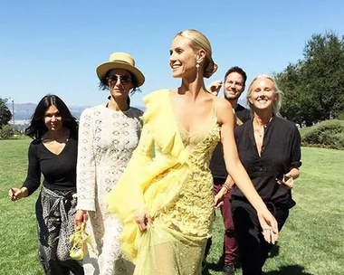 Woman in a yellow gown walks with a group on a sunny day in a grassy field.