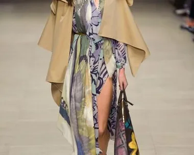 Model walks the runway in a floral dress with a slit, beige cape, and carries a patterned tote bag at Burberry's SS16 show.