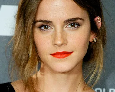 Emma Watson with light brown hair, wearing red lipstick and a black top, at a public event.