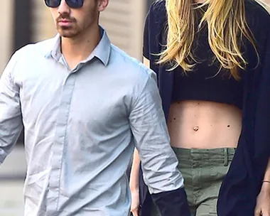 A man and a woman walking on a street. The man is in a gray shirt and sunglasses, the woman in a crop top and sunglasses.