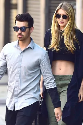 A man and a woman walking on a street. The man is in a gray shirt and sunglasses, the woman in a crop top and sunglasses.